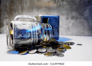 US Dollars And Coins Fill A Glass Tip Jar With Money.Looking Down On Glass Jar Filled With Coins On A Background Of Paper Money. Small Savings Add Up.Coins And Banknote In A Glass Jar 