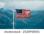 The USA American flag waving proudly with the Alaskan mountains in the background on a cloudy summer day