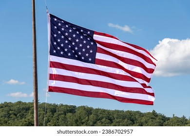 USA flag on flagpole flutters in the wind with red and white stripes and stars above a green forest and white clouded blue sky - Shutterstock ID 2386702957