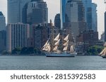 USCGC Eagle sails beneath Lower Manhattan skyscrapers on July 4. Vertical telephoto view places the tall ship below glass towers, brick buildings and waterfront trees.