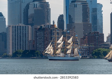 USCGC Eagle sails beneath Lower Manhattan skyscrapers on July 4. Vertical telephoto view places the tall ship below glass towers, brick buildings and waterfront trees.