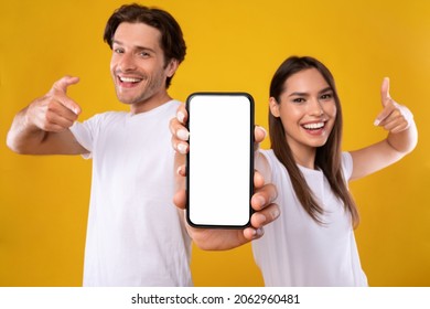 Use This New Application. Portrait Of Cheerful Millennial Couple Pointing At Smartphone With White Screen, Recommending Mobile App, Advertising Your Website, Mock Up For Design. Selective Focus