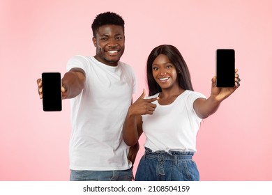 Use This New Application. Portrait of cheerful young black couple holding 2 cellphones in hands pointing at empty screen, recommending mobile app, advertising website, mock up for design, pink wall