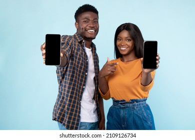 Use This New Application. Portrait Of Cheerful Young Black Couple Holding 2 Cellphones In Hands Pointing At Empty Screen, Recommending Mobile App, Advertising Website, Mock Up For Design, Blue Wall