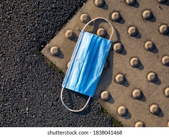 A Used, Blue Surgical Mask Used For COVID-19 PPE Protection, Discarded As Litter On A Pavement / Sidewalk In An Urban Area Causing Environmental Pollution