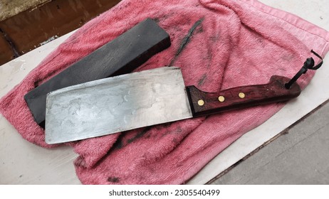 An used grindstone or whetstone and a sharp knife chopper with wooden handle laid together on an orange kitchen tower or rag