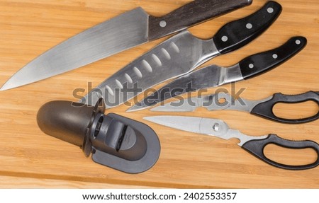 Used manual kitchen combined sharpener of broach type on a blurred background of the different old kitchen knives and scissors on the wooden cutting board, top view
