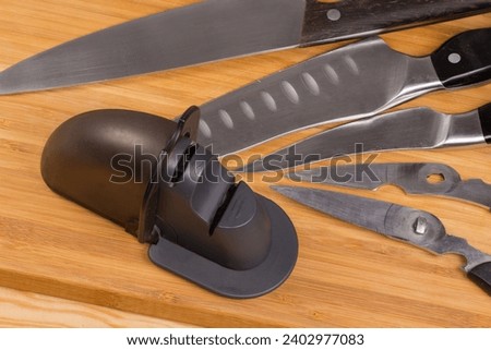 Used manual kitchen combined sharpener of broach type against the different old kitchen knives and two halves of scissors on the wooden cutting board
