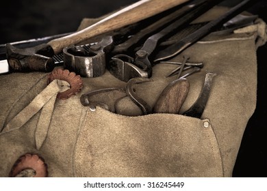 Used Tools of a farrier including rasp, hoof knife, chaps, hoof pick, and nippers draped over stool