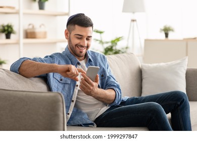 Useful App. Handsome Israeli Man In Kippah Using Smartphone While Relaxing On Couch At Home, Jewish Guy Sitting On Sofa In Living Room, Browsing Internet Or Messaging With Friends, Free Copy Space