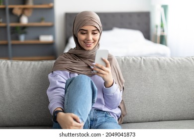 Useful App. Smiling Arab Lady In Headscarf Using Cell Phone Relaxing On Couch At Home, Middle Eastern Female Sitting On Sofa In Living Room, Browsing Internet Or Messaging With Friends, Free Space