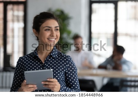 Useful technology. Happy friendly mixed race female manager consultant designer standing in modern office place with pleasant smile distracted from using tablet computer pc meeting client, copy space