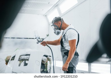 Uses painting gun. Caucasian automobile repairman in uniform works in garage.