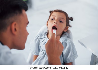 Uses Tonsil To Check Throat. Young Pediatrician Works With Little Female Visitor In The Clinic.