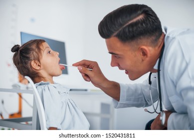 Uses Tonsil To Check Throat. Young Pediatrician Works With Little Female Visitor In The Clinic.