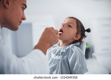 Uses Tonsil To Check Throat. Young Pediatrician Works With Little Female Visitor In The Clinic.