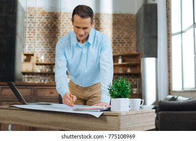 Using All Devices. Cheerful Male Person Looking Downwards While Drawing Project Work, Leaning On Table