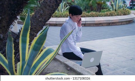 Using app on smartphone. Beautiful female working on the open air at the park with palm tree and exotic green bushes.On the background people sitting on the bench walking and riding cars.