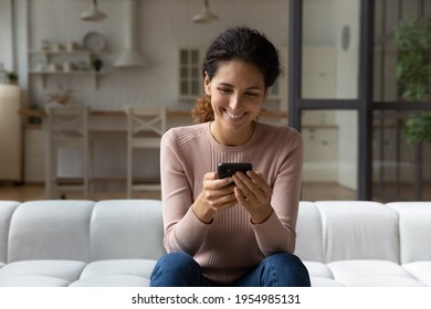 Using application. Smiling latin female spend free time sit on sofa look at smartphone screen chat at web app. Happy millennial woman enjoy communication online by cell play mobile game text message