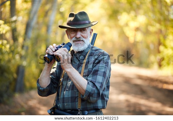 Using binoculars for better hunting on birds.\
Senior man holding binoculars during hunting in autumn forest look\
at camera. Road\
background