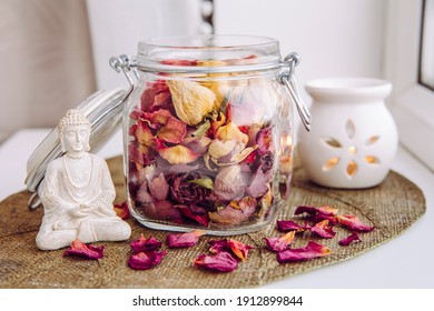 Using dry rose petals to make rose potpourri which is great for home scent. Mason jar filled with dry rose petals and puds with meditating Buddha figurine and aroma lamp with candle next to it.