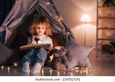 Using flashlight to read the book. Cute little girl is in the tent in domestic room.