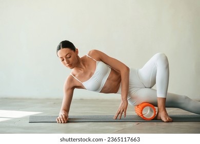 Using foam roller for exercises. Young woman with slim body type is in fitness clothes in the studio.