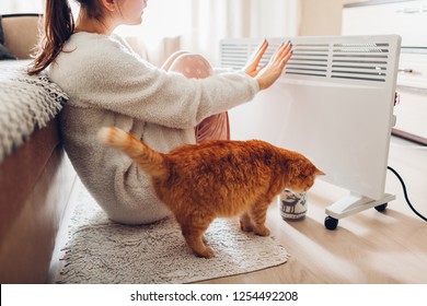 Using heater at home in winter. Woman warming her hands with cat. Heating season.