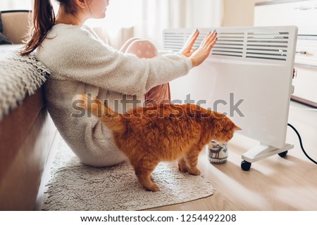Using heater at home in winter. Woman warming her hands with cat. Heating season.