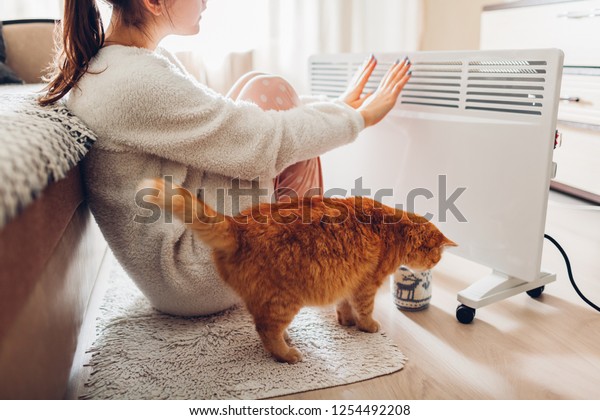 Using heater at home in winter. Woman warming her\
hands with cat. Heating\
season.
