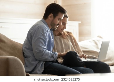 Using laptop. Concentrated young husband and wife sitting together on comfy sofa using laptop computer, making payments or purchases online, browsing internet sites, reading posts at social networks