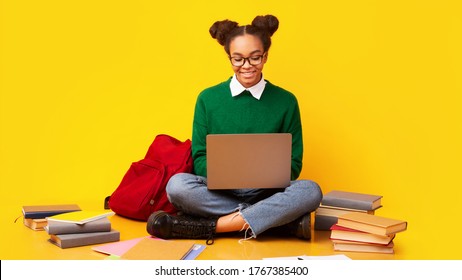 Using Laptop. Happy black teenage girl doing homework, sitting on the floor, yellow studio background, copyspace