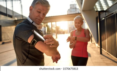 Using modern techologies. Active middle-aged couple in sports clothing checking training results while standing together outdoors. Checking pedometer