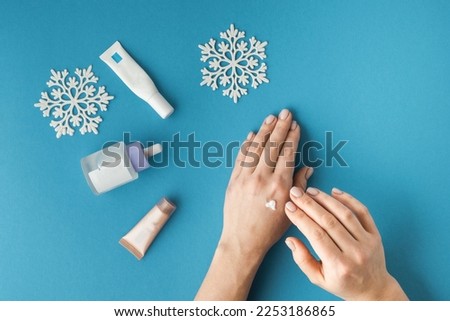 Using moisturizing hand cream during winter season. Female hands smearing white cream on the blue background next to different skin care products. Treatment for the skin advertisement