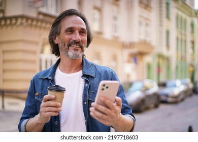 Using Navigation Mature Man Stand On Street Of European City Using Mobile Phone. Travel Concept. Handsome Middle Aged Man Enjoying His Coffee To Go Traveling In Old Town Wearing Casual. 