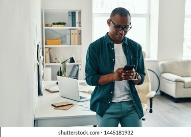 Using new mobile app. Handsome young African man in shirt using smart phone while standing in the office