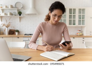 Using Online Open Sources. Confident Latina Woman Handwrite To Notepad Compare Data With Information At Web Pages. Young Lady Manage Organize Business Schedule Take Notes From Cellphone Screen By Hand