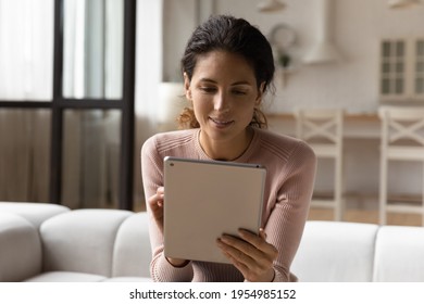 Using pad. Smiling latina woman user sit on couch at home surf web on tablet computer read ebook via wireless wifi internet. Happy young female enjoy online chatting with friend study language in app