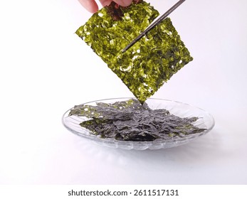 Using a pair of scissors, one hand cuts a sheet of crunchy nori or roasted seaweed. The objects are isolated on a white background. An eye-level shot.