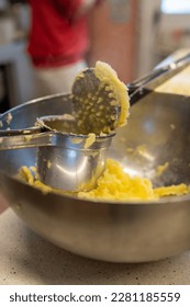 Using a potato ricer kitchen implement to mash boiled russet potatoes in an aluminum bowl