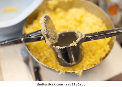 Using a potato ricer kitchen implement to mash boiled russet potatoes in a stainless steel bowl, close-up detail with soft-focus background