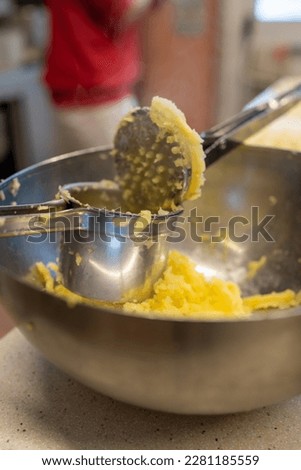 Using a potato ricer kitchen implement to mash boiled russet potatoes in an aluminum bowl