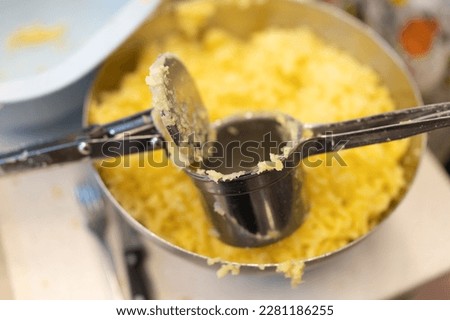 Using a potato ricer kitchen implement to mash boiled russet potatoes in a stainless steel bowl, close-up detail with soft-focus background
