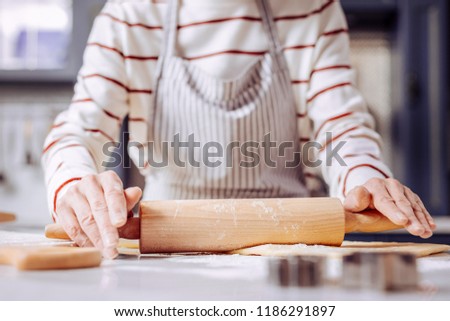 Using rolling pin. Careful enthusiastic aged woman standing next to the table and touching the rolling pin while making tasty cookies