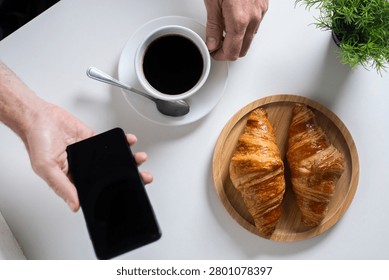 Using Smartphone During Breakfast with Coffee and Croissants on White Table. Top view