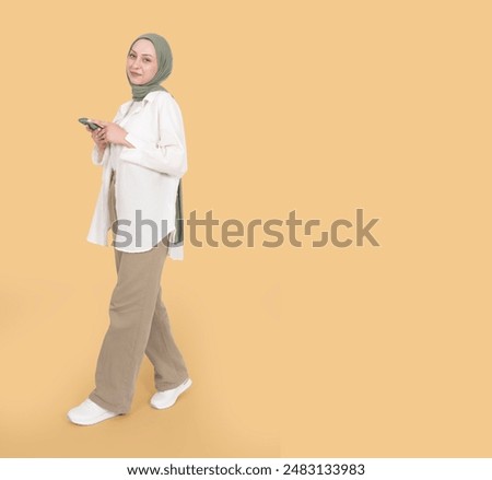 Using smartphone, full body view young caucasian muslim woman wear hijab abaya using smartphone. Walking holding mobile phone, looking camera over light orange beige background. Copy space.