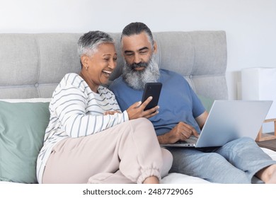 Using smartphone and laptop, senior couple relaxing on bed and smiling. Technology, communication, elderly, relaxation, lifestyle, bedroom - Powered by Shutterstock