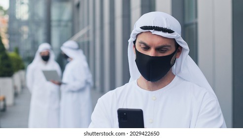 Using smartphone. Waist up portrait view of the arabic businessman wearing protective mask writing serious message to somebody while holding his cell phone at the street. Pandemic concept