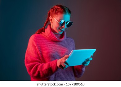 Using tablet  Caucasian woman's portrait isolated gradient studio background in neon light  Beautiful female model and sunglasses  red hair  Concept human emotions  facial expression  ad 