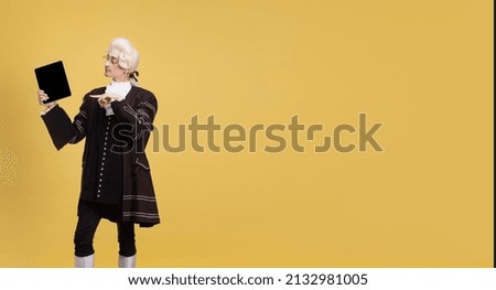 Using tablet. Portrait of young elegant man in white wig and vintage medieval outfit posing isolated on yellow background. Art, beauty, fashion. Retro style, eras comparison. Copy space for ad